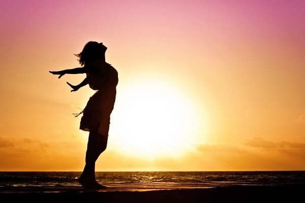 A woman stands silhouetted by the sea, in front of an amazing pink and orange sunset. She's throwing her arms and head back as if celebrating her freedom.