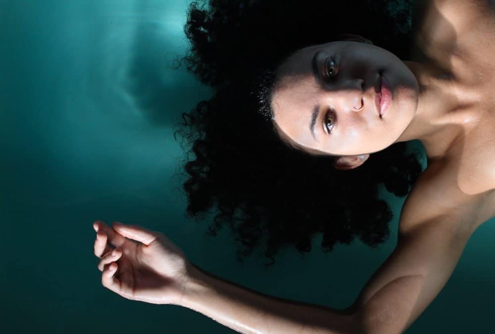 A young lady with curly dark hair floats on her back in water, the photo is taken from above.